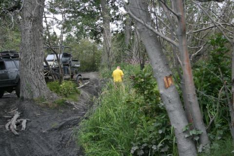 1990 Chevrolet Suburban - A dark-colored SUV is parked on a muddy, off-road trail next to a larger tree. A person in a yellow shirt is walking away from the vehicle…