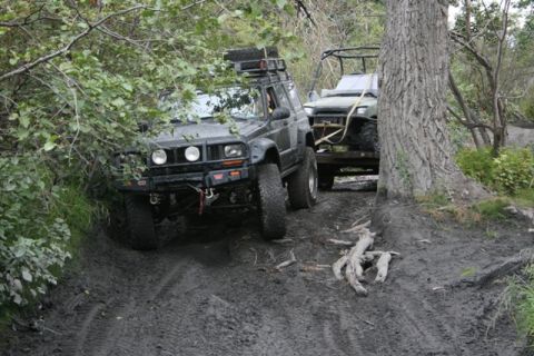 1990 Chevrolet Suburban - A black Jeep Cherokee is driving through a muddy trail, pulling a trailer with a green ATV on it. The vehicle is surrounded by trees and fo…