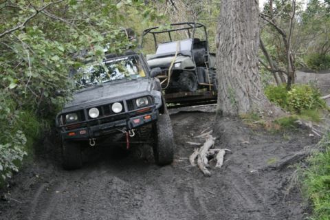 1990 Chevrolet Suburban - A dark-colored Jeep Cherokee is navigating a muddy, off-road trail, pulling a trailer with a green ATV. The vehicle is surrounded by dense…