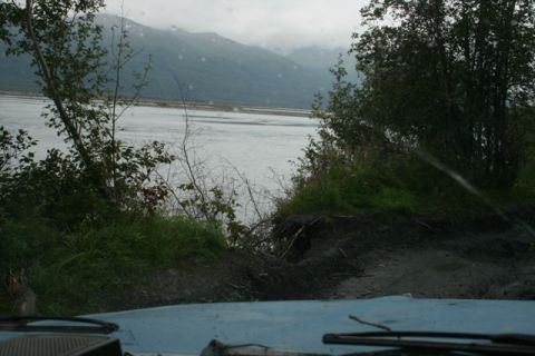 1990 Chevrolet Suburban - View from the driver's seat of a light blue vehicle looking out at a wide, silty river and mountains under a cloudy sky. The foreground sho…
