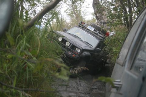 1990 Chevrolet Suburban - A black Jeep Cherokee is stuck in thick mud, tilted at an angle with its front tires deep in the mire. The vehicle has a roof rack with a s…
