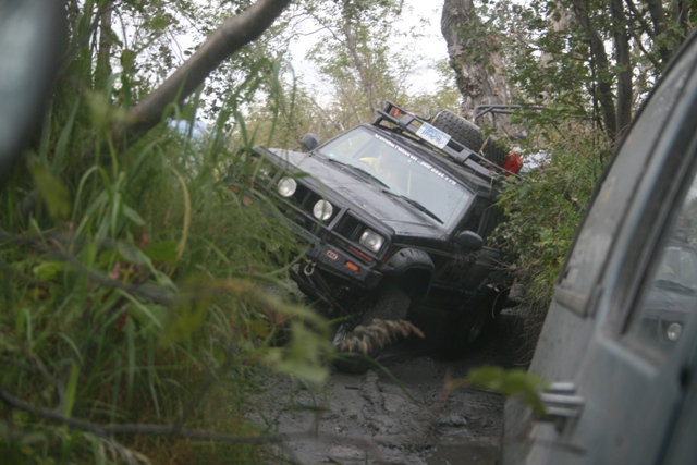 Black Jeep Cherokee stuck in mud, tilted, with roof rack and spare tire, surrounded by trees and foliage.