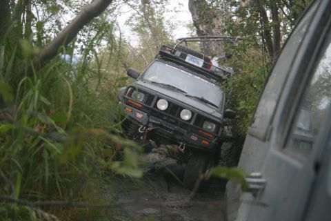1990 Chevrolet Suburban - A black Jeep Cherokee is navigating a muddy, overgrown trail, with another vehicle partially visible on the right. The Jeep is equipped wit…