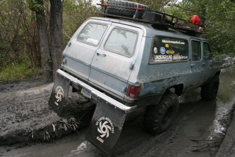 1990 Chevrolet Suburban - A light blue Chevrolet Suburban is navigating a muddy, off-road trail, with its rear mud flaps prominently displaying logos. The vehicle is…