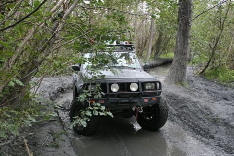 1990 Chevrolet Suburban - A dark-colored SUV with a bull bar and roof rack navigates a muddy, wooded trail with standing water.