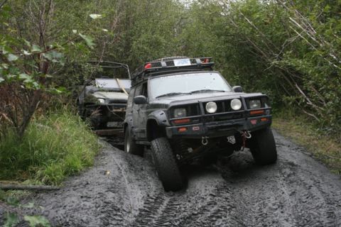 1990 Chevrolet Suburban - A dark-colored Jeep Cherokee XJ is navigating a muddy trail, with its front tires deep in a rut. Another off-road vehicle is visible behind…