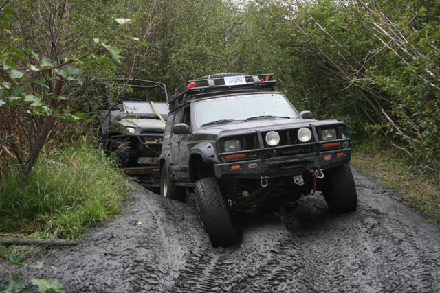 Black Jeep Cherokee XJ on a muddy trail with another vehicle behind it.