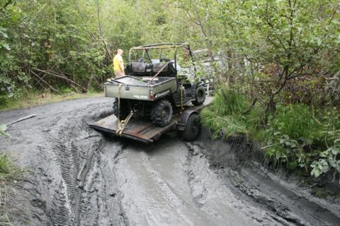 1990 Chevrolet Suburban - A green Polaris Ranger utility vehicle is on a trailer stuck in deep mud on a trail. A person in a yellow shirt stands nearby.