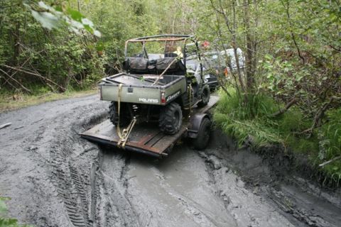 1990 Chevrolet Suburban - A green Polaris Ranger utility vehicle is on a trailer stuck in a muddy, wooded trail.