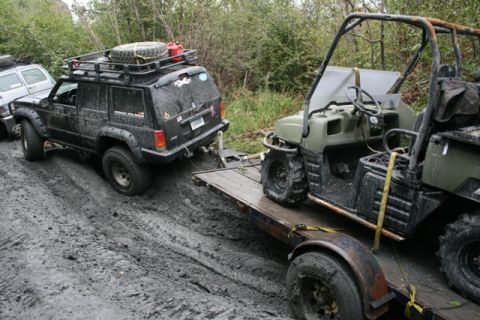 1990 Chevrolet Suburban - A black Jeep Cherokee is being towed out of thick mud by a utility vehicle on a trailer. The Jeep has a roof rack with a spare tire and gas…