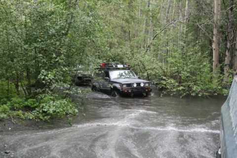 1990 Chevrolet Suburban - Two black SUVs are driving through a shallow, muddy creek surrounded by dense green foliage. The lead SUV has a roof rack with gear and is…