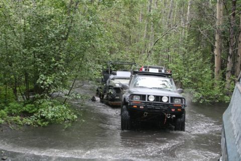 1990 Chevrolet Suburban - A dark-colored Jeep Cherokee is driving through a shallow, muddy stream in a wooded area. It is towing a trailer with an ATV on it.