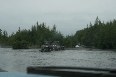 1990 Chevrolet Suburban - Two dark-colored SUVs are driving through a shallow body of water surrounded by trees. A third vehicle, possibly a boat, is visible further…