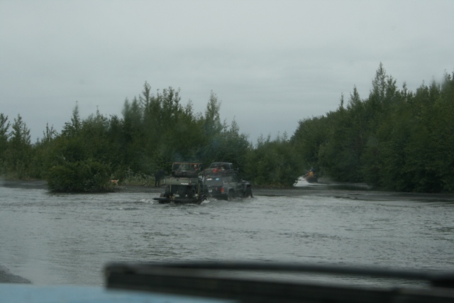 Two SUVs driving through water with trees on both sides.