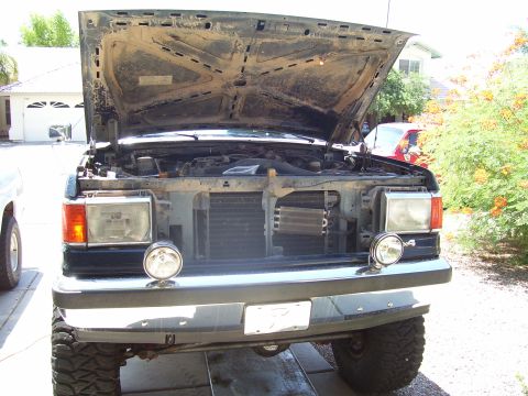 1991 Ford Bronco - The front of a dark-colored Ford Bronco with its hood open, revealing the engine bay. The vehicle has large off-road tires and aftermarket…