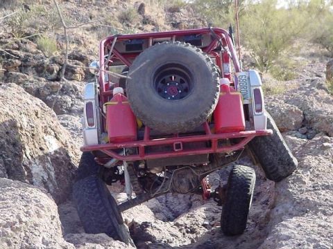 1991 Ford Bronco - Rear view of a red full-size Bronco-style rock crawler flexing over boulders on a desert trail, with the body cut away to expose a red roll…