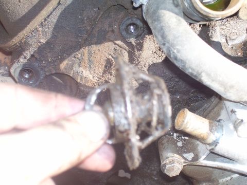 1991 Ford Bronco - A hand holds a rusted and broken thermostat housing in front of a dirty engine block. A hose and other engine components are visible in the…