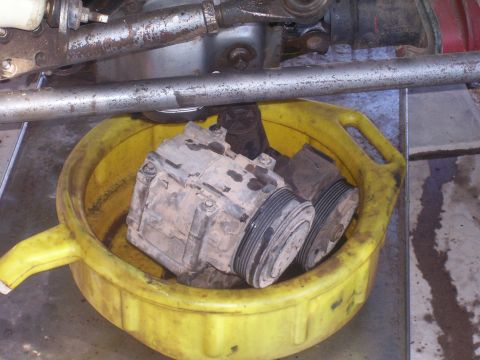 1991 Ford Bronco - Two dusty automotive AC compressors sit in a yellow plastic drain pan, with vehicle suspension parts visible above. The compressors appear…