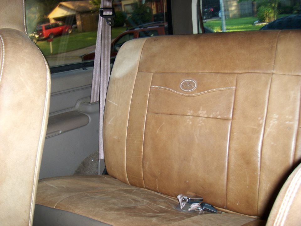 Interior of a vehicle with tan leather seats and a seatbelt, with a red SUV visible through the window.