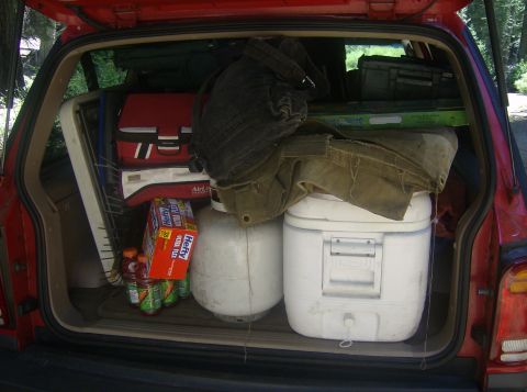 1986 Ford Bronco - The open rear cargo area of a red SUV is packed with camping gear, including coolers, bags, and bottles.