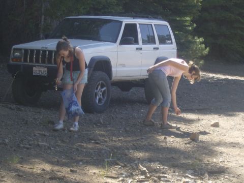 1986 Ford Bronco - Two young women are near a white Jeep Cherokee SUV parked on a dirt road. One woman is holding a blue bag and keys, while the other is bend…