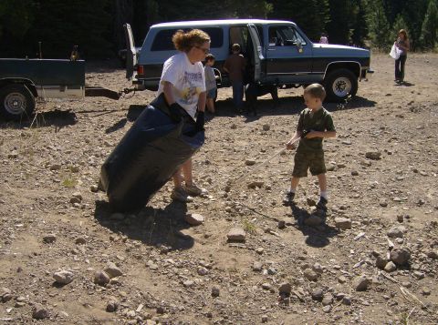 1986 Ford Bronco - A woman carries a large black trash bag while a young boy uses a grabber tool to pick up litter near a vintage SUV and a trailer.
