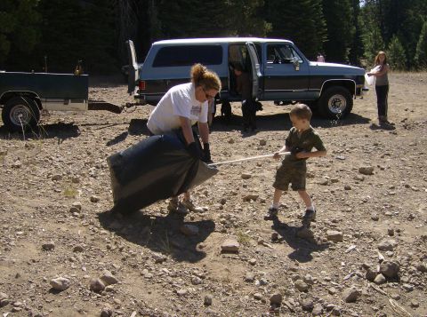 1986 Ford Bronco - A woman and a young boy are cleaning up trash near a blue SUV. The woman is holding a large black trash bag, and the boy is holding a pole…