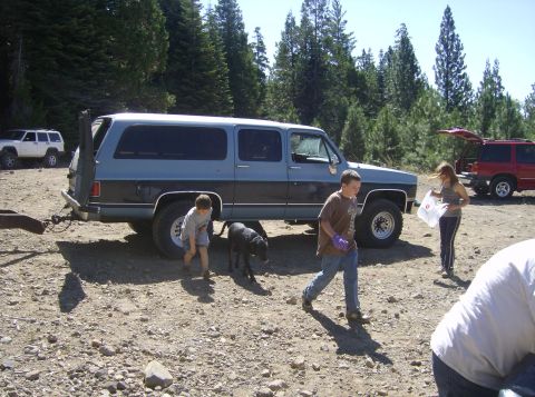 1986 Ford Bronco - A light blue and dark blue two-tone SUV is parked on a dirt lot with trees in the background. Several children and a black dog are near the…