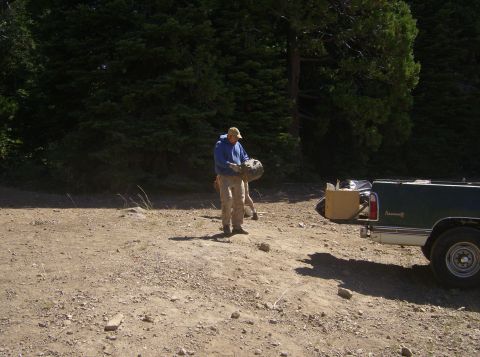 1986 Ford Bronco - A man in a blue hoodie and tan pants holds a large, round object while standing in a dirt lot next to a dark green pickup truck. The truck…