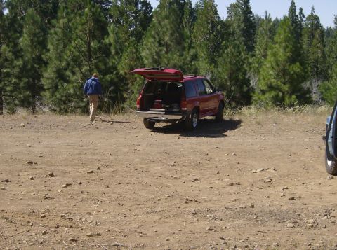 1986 Ford Bronco - A person walks away from a red SUV with its rear hatch open, parked on a dirt road in a wooded area.
