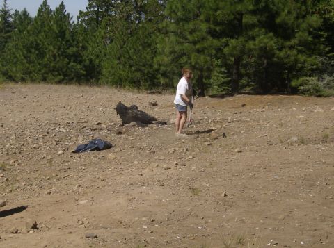 1986 Ford Bronco - A woman in a white t-shirt and denim shorts is raking the dry, rocky ground in front of a line of pine trees. A dark blue jacket or bag is…