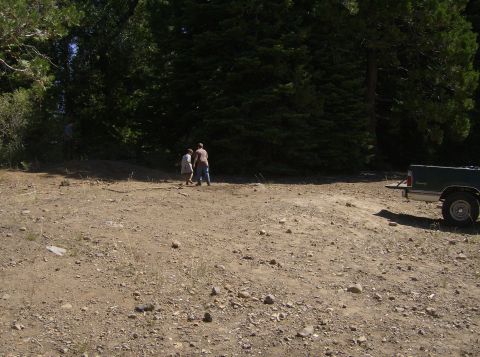 1986 Ford Bronco - A dark green pickup truck is parked on a dirt lot with two people standing in the background near a wooded area.