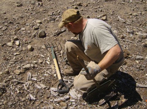 1986 Ford Bronco - A man wearing a cap and gloves is kneeling on a rocky, debris-strewn ground. He is holding a rake and a pitchfork is lying next to him.