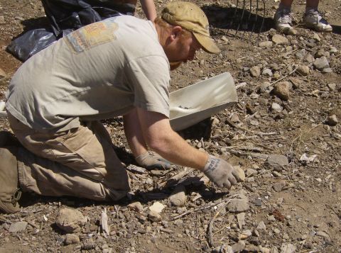 1986 Ford Bronco - A person wearing a tan t-shirt and khaki pants is kneeling on the ground, examining rocks and debris with a small white scoop nearby. They…