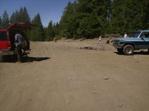 1986 Ford Bronco - A blue pickup truck is parked on a dirt lot with trees in the background. A red SUV is parked to the left with its rear hatch open.