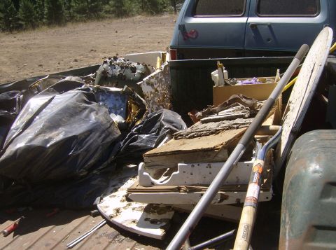 1986 Ford Bronco - The bed of a pickup truck is filled with debris, including a large black trash bag, broken wood, and metal pieces. A portion of a blue van…