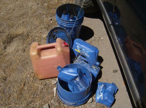 1986 Ford Bronco - Several blue plastic oil containers and a red plastic gasoline jug are scattered on dry grass and dirt next to a vehicle's tire and dark-co…