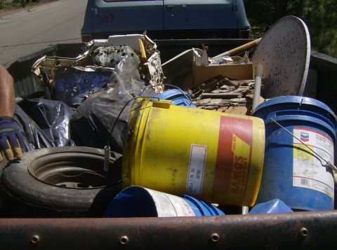 1986 Ford Bronco - The bed of a pickup truck is filled with various items including a tire, large plastic buckets, and debris. A blue van is visible in the ba…
