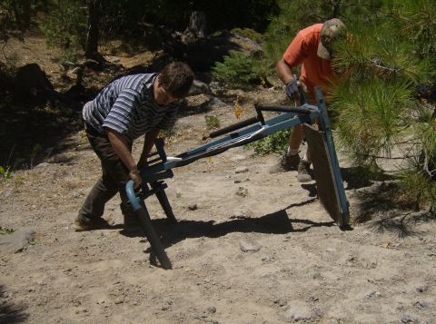 1986 Ford Bronco - Two people are working on a blue and black metal contraption on a dirt path in a wooded area. One person is wearing a striped shirt and the…