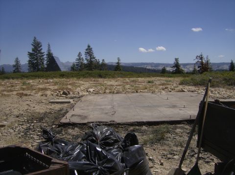 1986 Ford Bronco - A concrete slab foundation sits in a dry, rocky landscape with trees and mountains in the background. Several black trash bags are piled in…