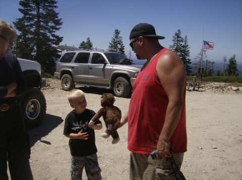 1986 Ford Bronco - A man in a red tank top holds a stuffed monkey for a young boy in a black t-shirt standing in front of a silver SUV.