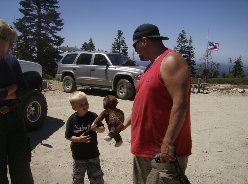 Man in red tank top holding stuffed monkey for a boy in black t-shirt, silver SUV behind them.