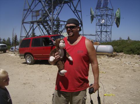 1986 Ford Bronco - A man in a red tank top holds a stuffed monkey in front of a red Ford Explorer SUV parked on a dirt lot next to a tall metal tower.