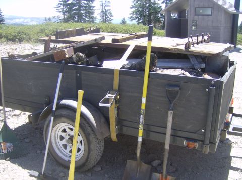 1986 Ford Bronco - A utility trailer loaded with debris and tools is parked in front of a rustic outhouse. Tools visible include a rake, two shovels, and a br…