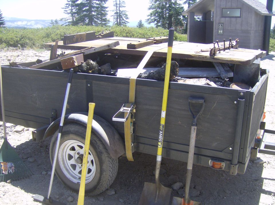 Utility trailer loaded with debris and tools, parked in front of an outhouse.