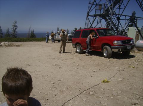 1986 Ford Bronco - A red Ford Explorer SUV is parked on a dirt lot next to a tall metal tower. A woman is leaning into the driver's side of the SUV.
