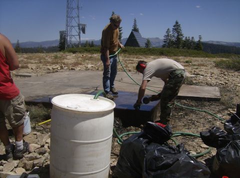 1986 Ford Bronco - Two men are working outdoors on a concrete pad near a large white barrel and black trash bags. One man is holding a green hose, and the oth…