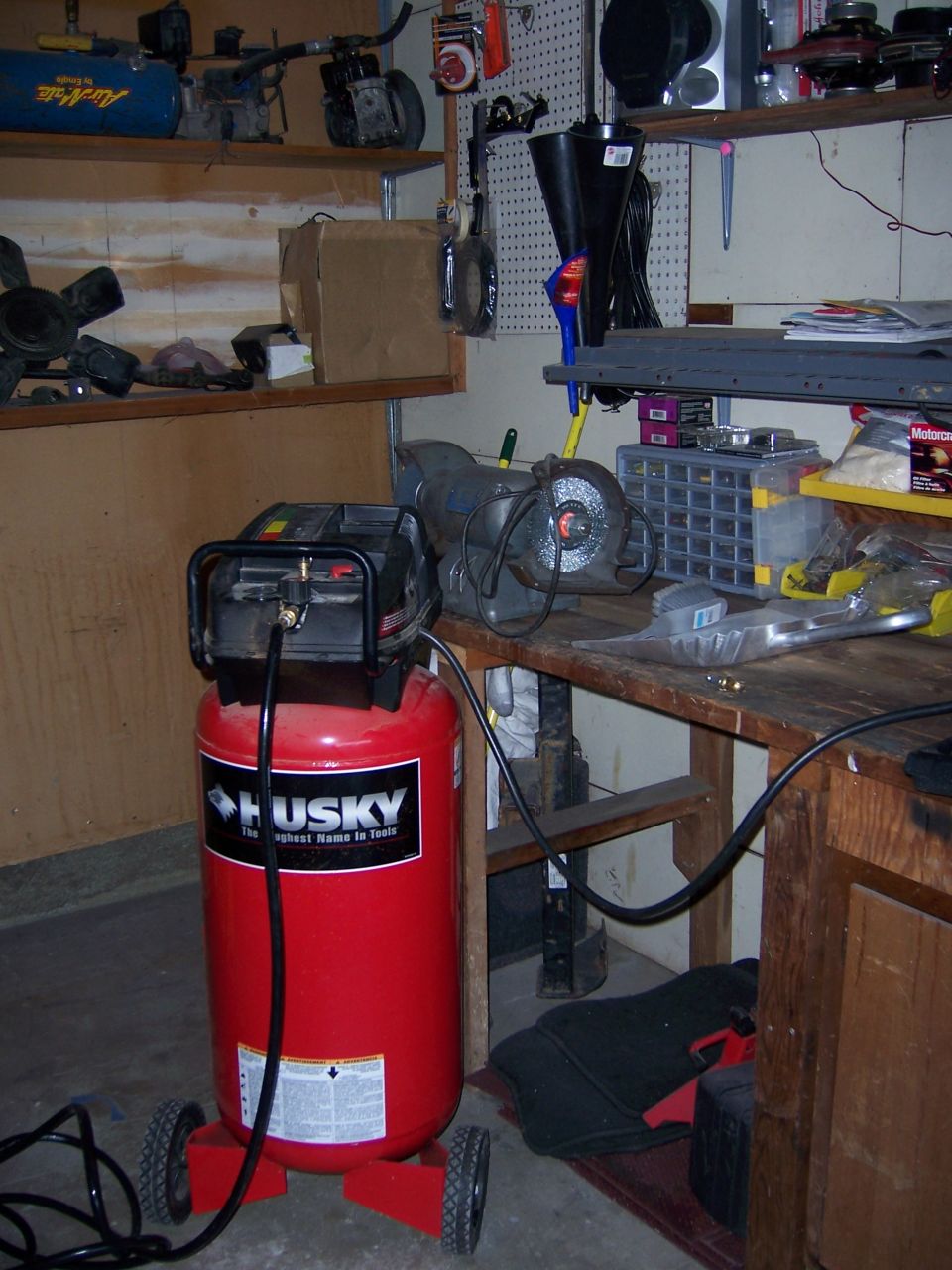 Red Husky air compressor with black hoses on a concrete floor next to a wooden workbench with tools