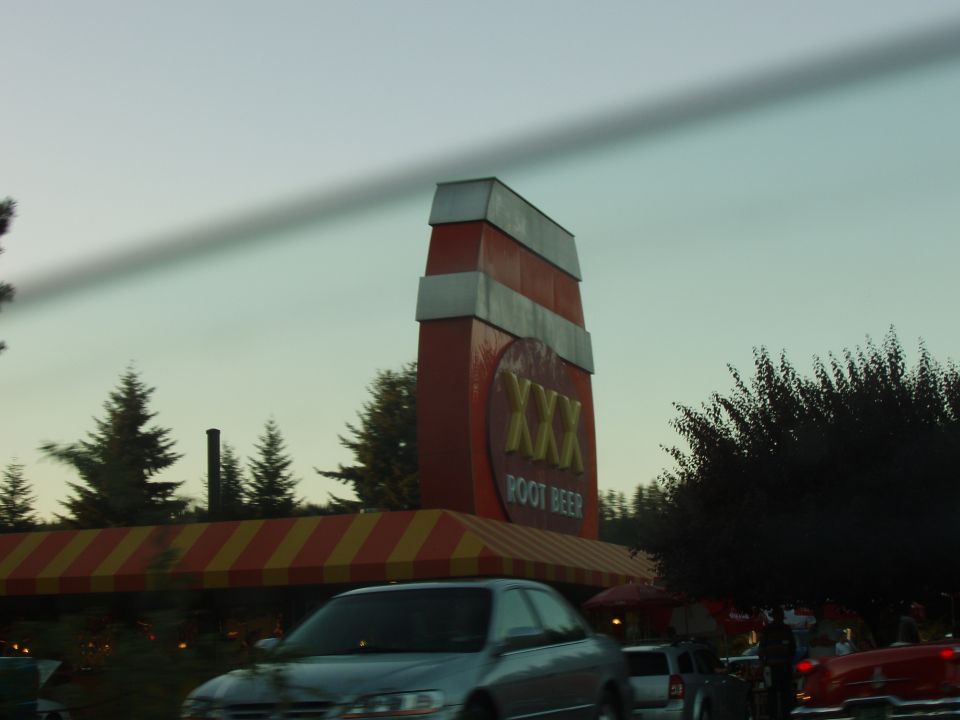 Silver sedan driving past a tall XXX Root Beer sign with a striped awning
