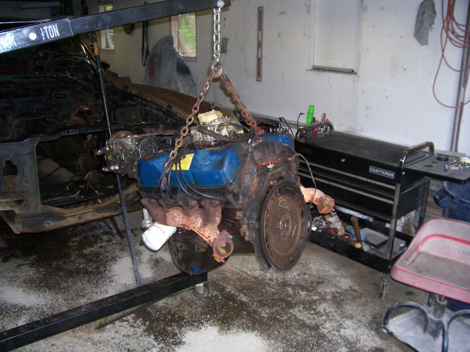 Rusty Ford V8 engine with blue valve covers hanging from hoist in a garage